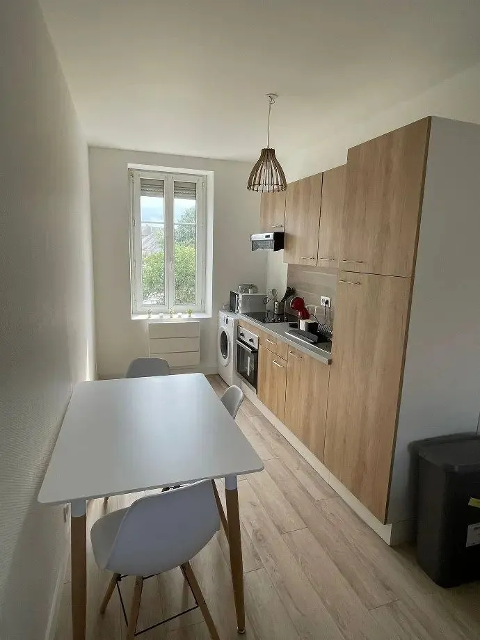 Cuisine équipée bois clair avec plaque, four et table de repas près de la fenêtre.