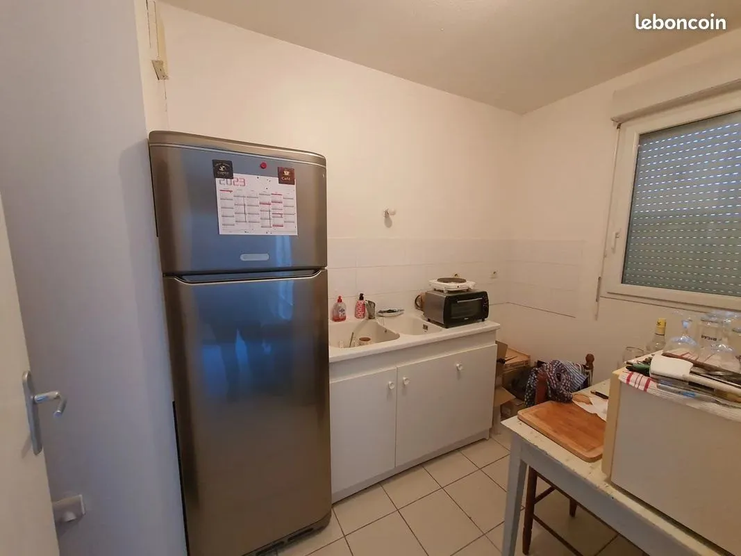 Cuisine d'origine avec réfrigérateur, plan de travail encombré et carrelage au sol.