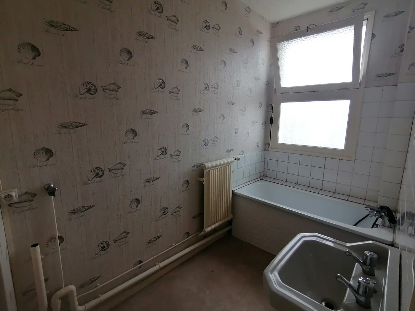 Salle de bains d'origine : papier peint à motifs, baignoire et carrelage blanc.