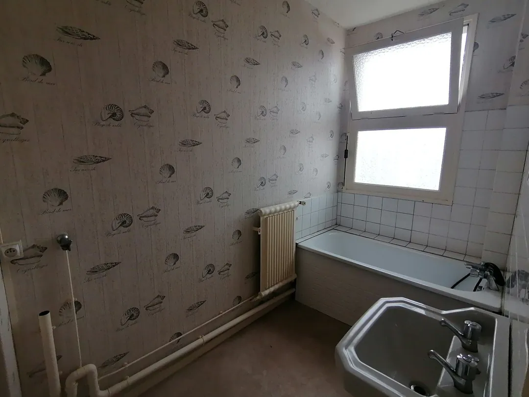 Salle de bains d'origine : papier peint à motifs, baignoire et carrelage blanc.