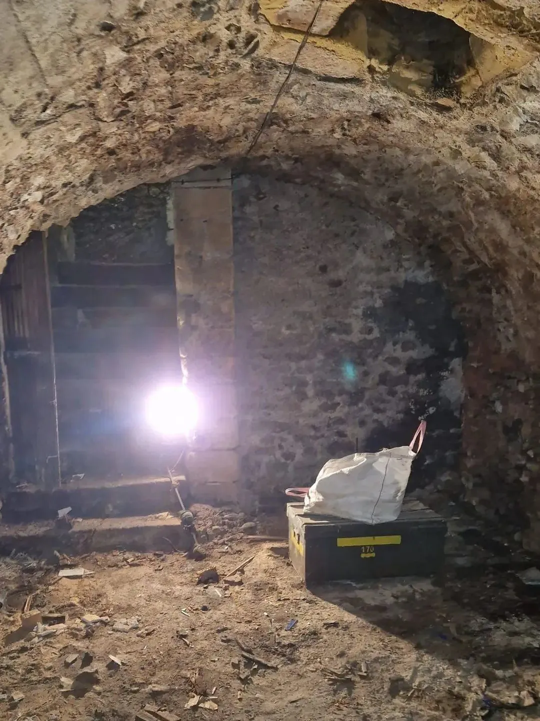 La même cave voûtée éclairée par un projecteur de chantier, sol en terre.
