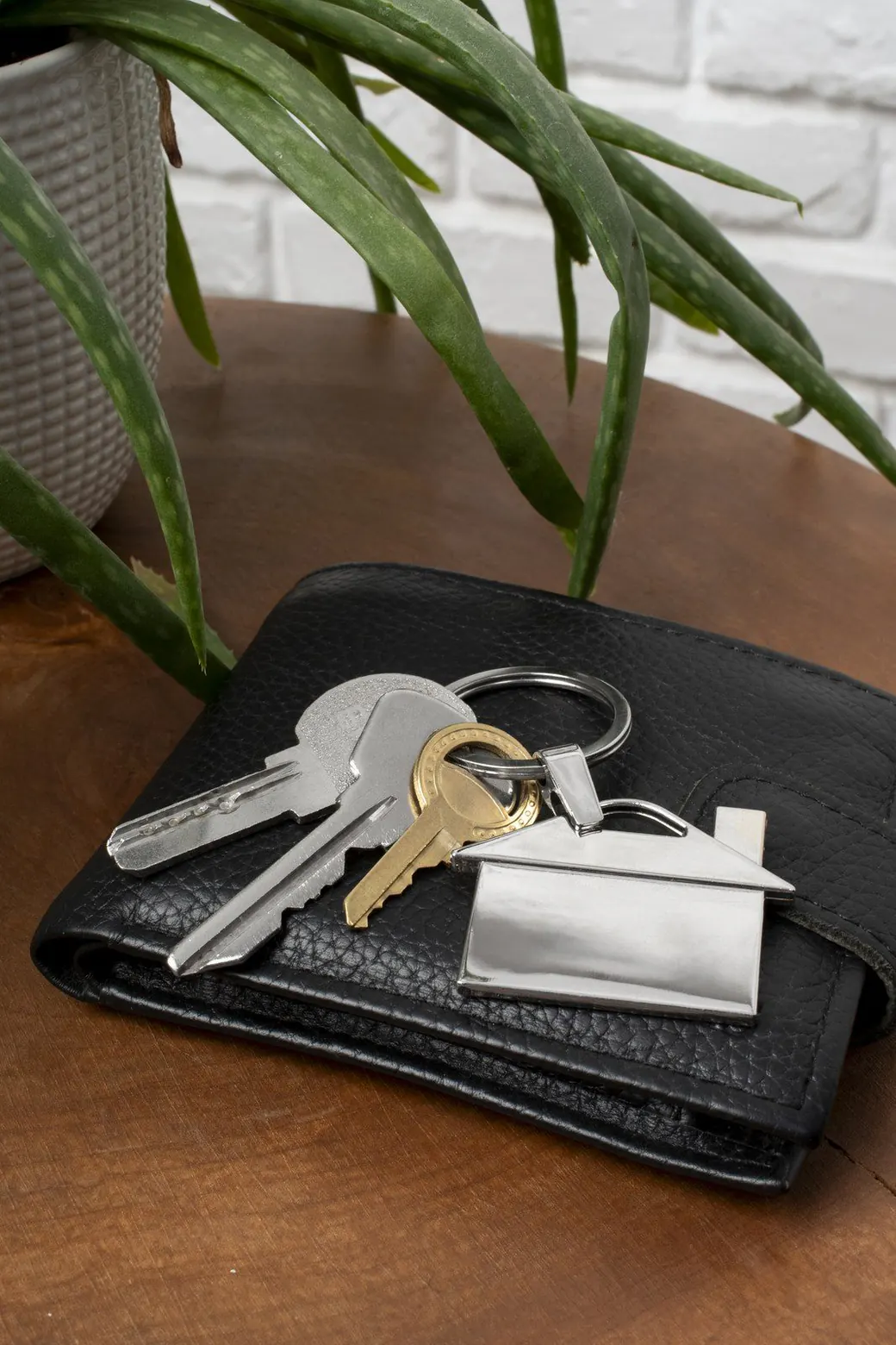 Un trousseau de clés avec un porte-clés en forme de maison, posé sur un portefeuille en cuir noir.
