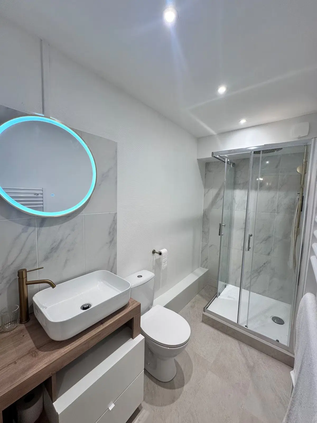 Salle d'eau neuve : douche vitrée, vasque sur plan bois et miroir rond.