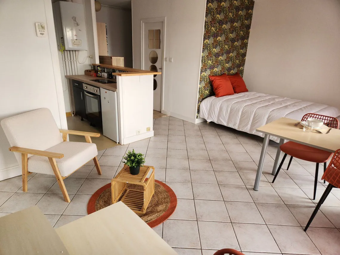 Studio meublé : lit, fauteuils, table basse et cuisine ouverte au fond.