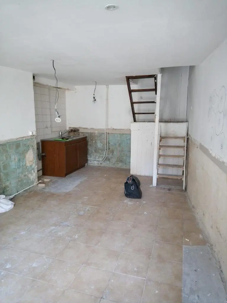 Pièce vide avec escalier de meunier, meuble bas ancien et sol brut.