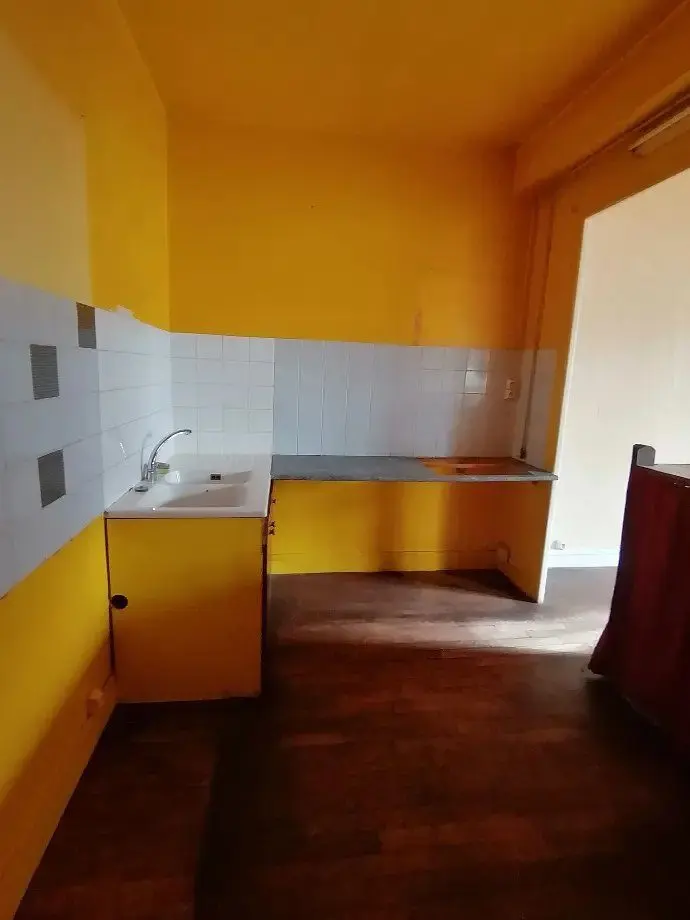 Cuisine d'origine aux murs jaunes, évier ancien et sol usé.