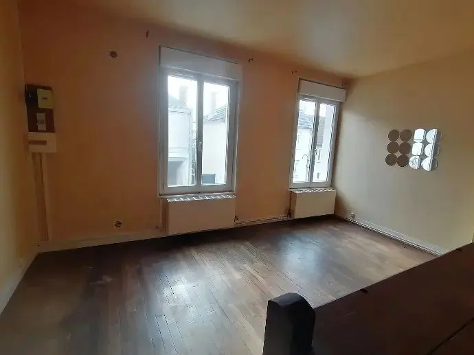 Séjour vide au parquet ancien, deux fenêtres et murs clairs.
