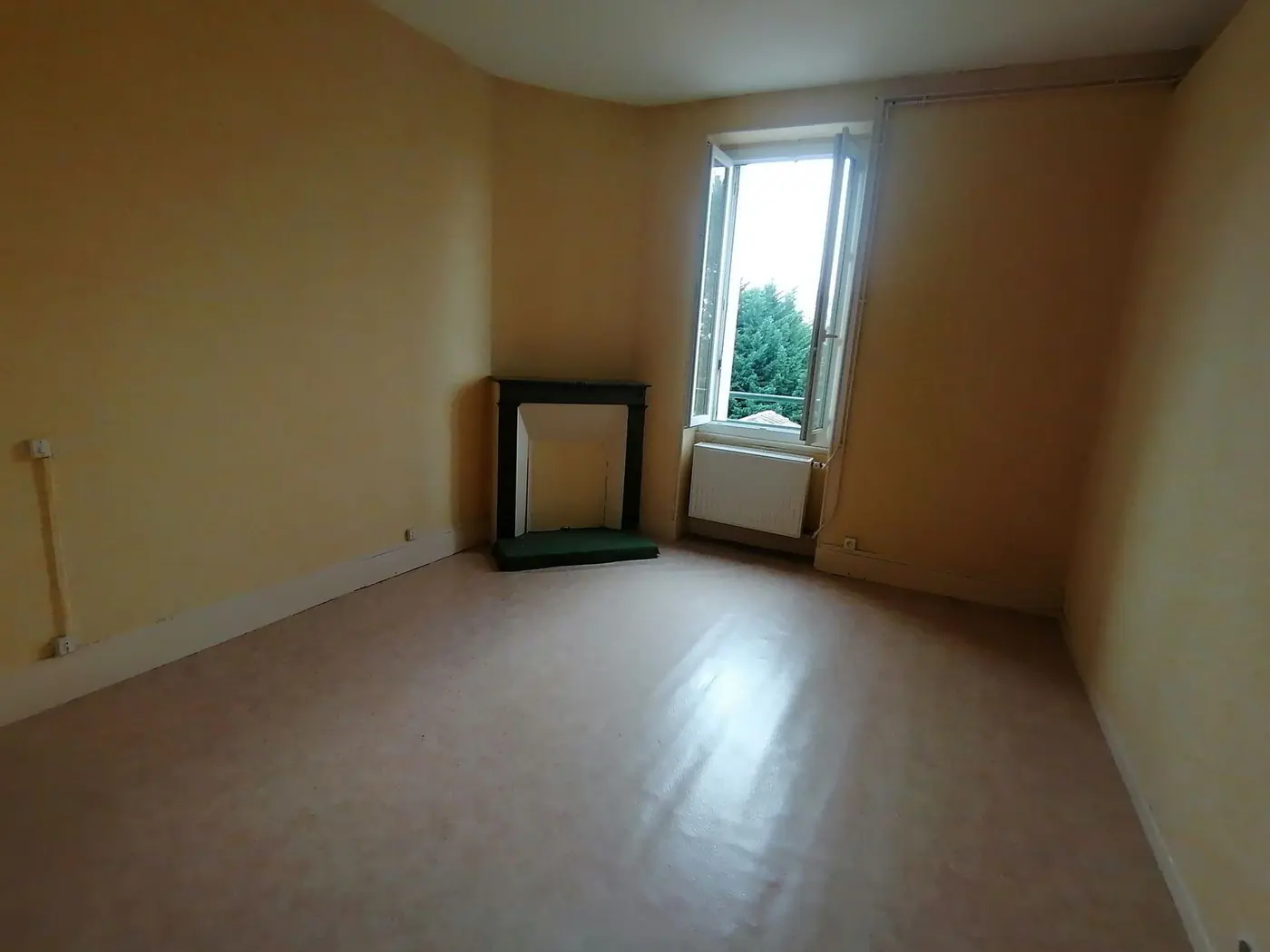 Salon vide aux murs jaunis, cheminée condamnée et fenêtre.
