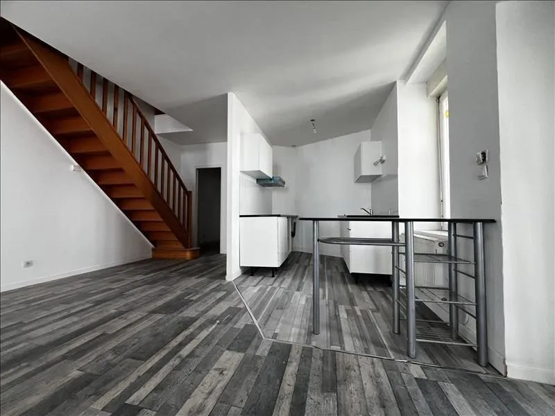 Séjour au sol stratifié gris avec escalier repeint et cuisine au fond.
