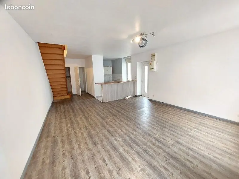 Séjour vide au parquet clair avec escalier en bois au fond.