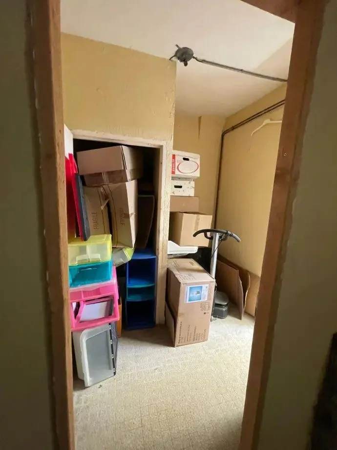 Ancienne réserve encombrée de cartons, de bacs de rangement et de matériel.