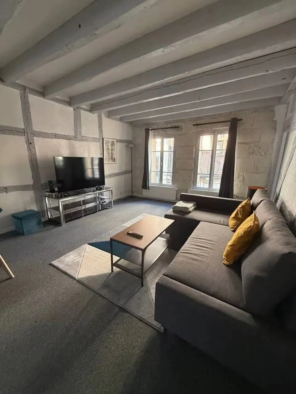Séjour du T4 : canapé d'angle, table basse, télévision, poutres blanches au plafond.
