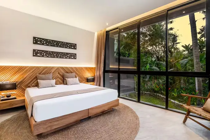 Une chambre de la villa, tête de lit en bois et baie vitrée sur la végétation.